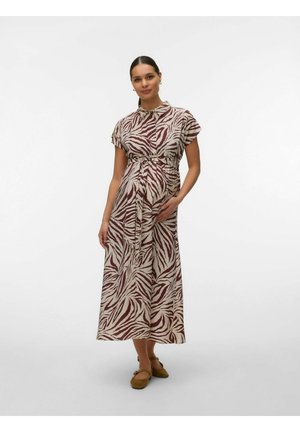 Femme enceinte aux cheveux foncés portant une robe zèbre beige et marron et des chaussures plates marron, debout avec une main sur son ventre.