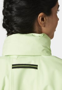 Chaqueta impermeable verde claro con un alto cuello, que presenta una franja negra y reflectante en horizontal en la parte posterior. Textura suave, con un diseño minimalista.