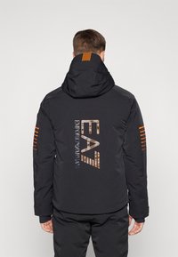 Čierna puffa bunda s oranžovými pruhmi na rukávoch a chrbte. Obsahuje kapucňu a potlač loga na zadnej strane. Hladká textúra, odolný materiál.