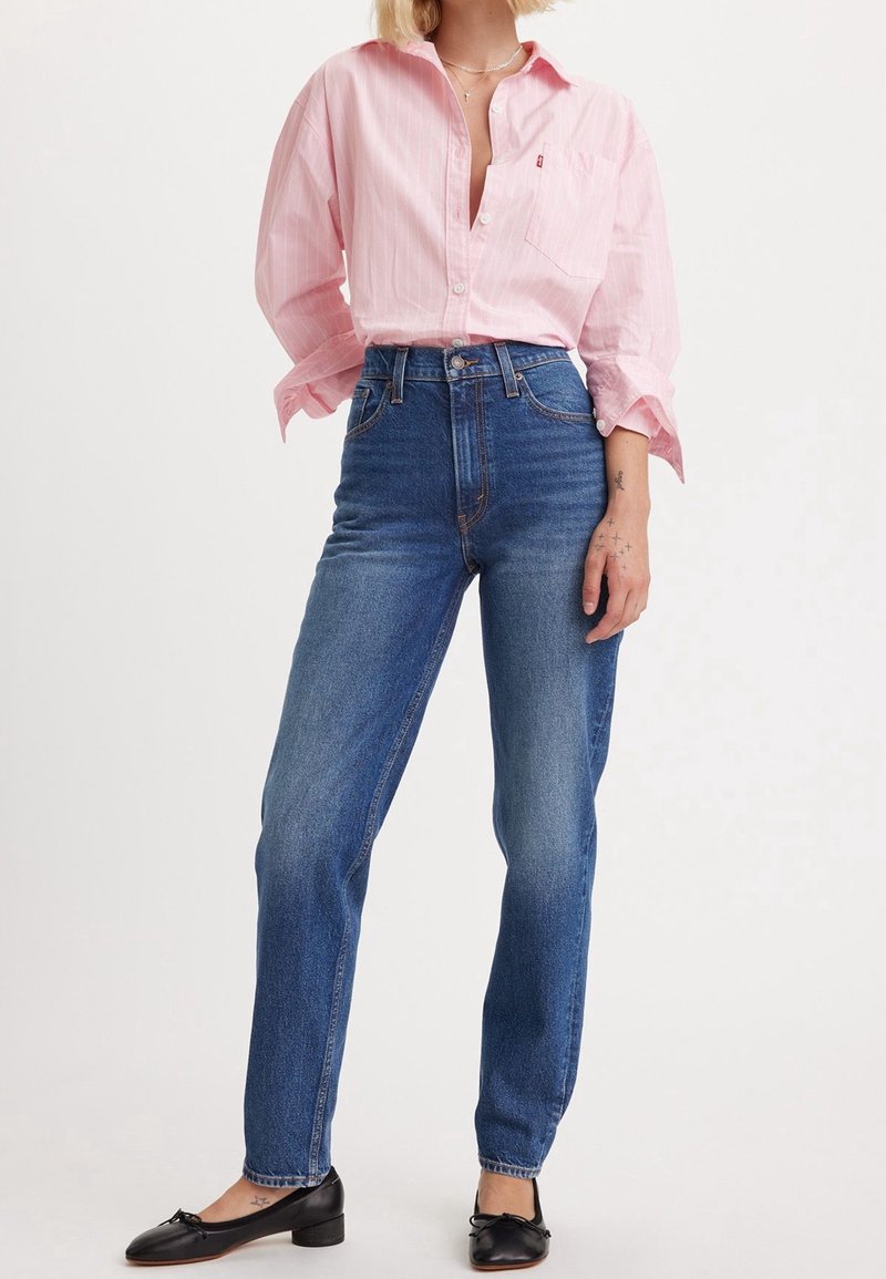 Personne portant un jean bleu taille haute, une chemise rose clair à boutons avec les manches retroussées, des ballerines noires et peu de bijoux sur un fond uni.