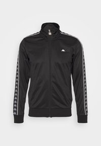 Chaqueta deportiva negra con cremallera hecha de tela suave, que presenta un collar alto, franjas laterales y un pequeño logo en el lado izquierdo del pecho.