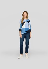Blaues Oberteil mit geometrischem Muster und Dreiviertelärmeln, kombiniert mit dunkelblauen Jeans und weißen Sneakers. Weiche Textur, lässiges Design.