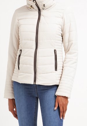 Woman wearing a light beige quilted jacket with front zipper and zipped pockets, paired with blue jeans against a white background.
