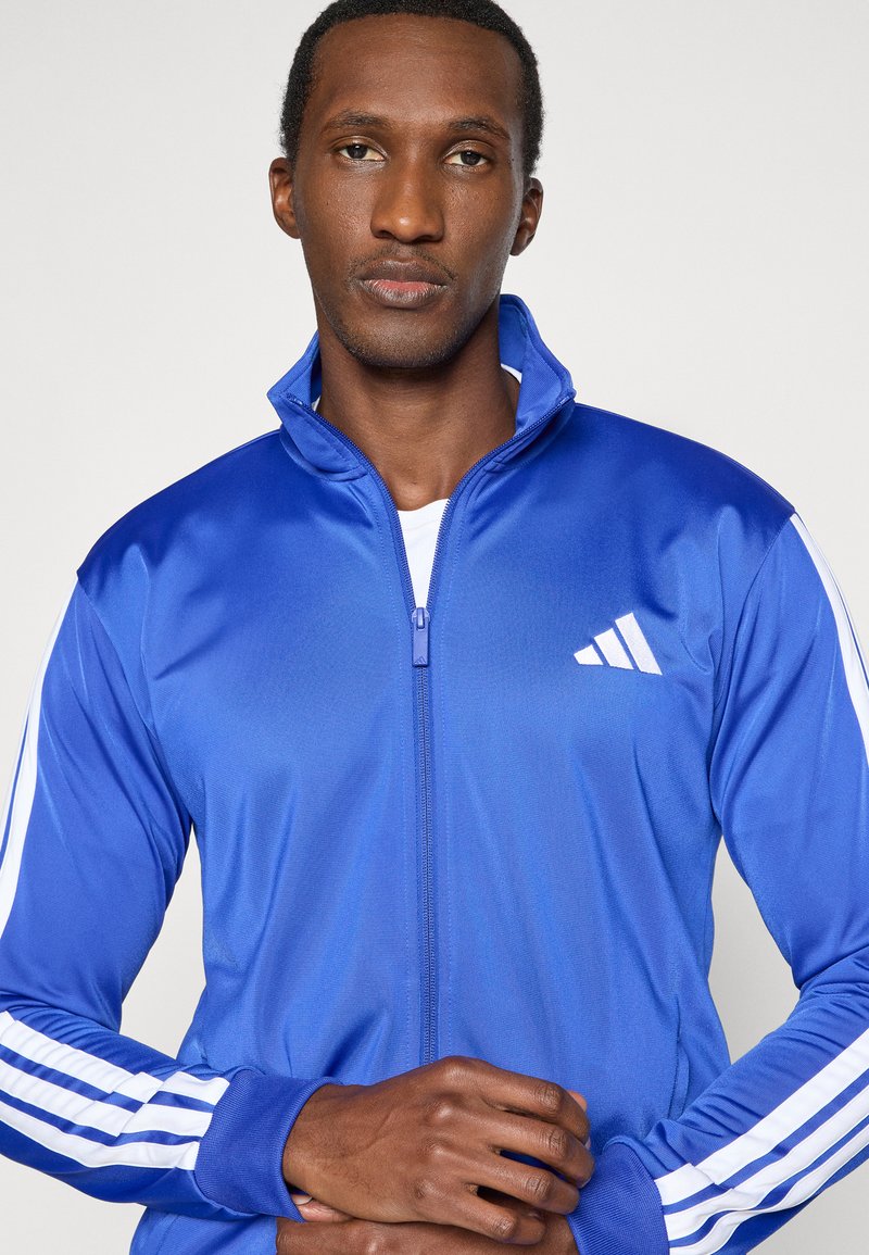 Blaue Sportjacke aus glattem Stoff. Mit hohem Kragen, durchgehendem Reißverschluss und weißen Streifen an den Ärmeln. Markenlogo auf der Brust.