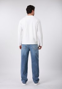 Άσπρο πουκάμισο με μακριά μανίκια, συνδυασμένο με ανοιχτό μπλε, φαρδιά τζιν παντελόνια, που παρουσιάζουν λεπτομερείς ραφές στην πλάτη και κλασική υφή denim.