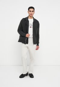 Fekete bőr keresztpántos táska, szürke kötött kardigán, fehér póló logóval, világos színű nadrág és fekete öltönycipő.