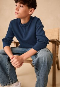 Tumši zils ribots krekls ar garām piedurknēm un Henlija apkakli, kopā ar gaisīši zilām, brīva piegriezuma džinsu biksēm. Fona redzama koka krēsls.