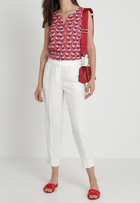 Rotes und blau gemustertes ärmelloses Oberteil, weiße maßgeschneiderte Hose, rote flache Sandalen und eine kleine rote Handtasche mit langem Tragegurt.