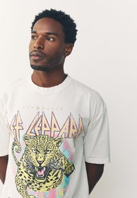White cotton t-shirt features a graphic of a roaring leopard in yellow, with multicolored text "Def Leppard" and abstract accents.