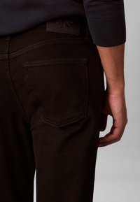 Κοντινό σε γοφό και χέρι ατόμου που φοράει μαύρο τζιν με ορατή πίσω τσέπη και μαύρη δερμάτινη ετικέτα Calvin Klein.