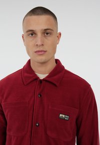 Rode fleece jas met een kraag, twee borstzakken en een klein logo-label. Beschikt over zwarte knopen en een zachte, gestructureerde oppervlakte.