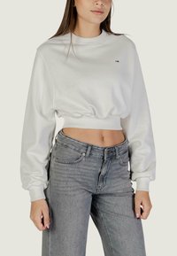 Felpa bianca corta con maniche lunghe, polsini a coste e un piccolo logo sul petto, abbinata a jeans grigi a vita alta.