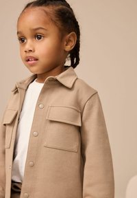 Jeune enfant avec des cheveux tressés portant une veste beige à boutons sur une chemise blanche, regardant légèrement vers la gauche sur un fond beige.