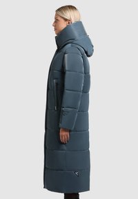 Pikk, tume sinine puhvkattega jakk kõrge kraega ja kapuutsiga. Omab tikkimist sisaldavaid sektsioone, lukuga küljetaskuid ja logotähtsust hemmi lähedal.