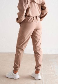 Survêtement léger beige avec taille élastique et poignets élastiques. Il présente une coupe décontractée et est associé à des chaussures de sport blanches.