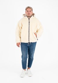 Crème kleurige fleecejack met een capuchon, ritssluiting aan de voorkant en twee zijzakken. Draagt met blauwe jeans en witte sneakers.