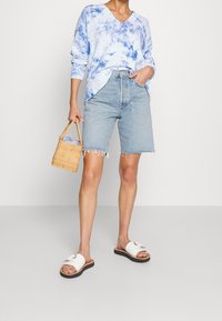 Ljusblå tie-dye tröja, högmidjade jeansshorts med fransad nederkant och vita slidesandaler. Accessoarer: liten vävd handväska.