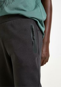 Mørkegrå joggingbukser med en glat tekstur, der har en lynlåslomme og en afslappet pasform, kombineret med en skjorte i tealgrøn.