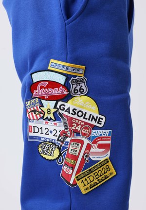 Tissu bleu avec des patchs colorés présentant des enseignes de station-service vintage, une pompe à essence, un bouclier Route 66 et des plaques d'immatriculation du Montana et du Colorado.