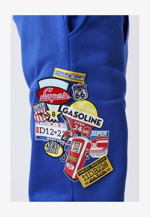 Tissu bleu avec des patchs colorés présentant des enseignes de station-service vintage, une pompe à essence, un bouclier Route 66 et des plaques d'immatriculation du Montana et du Colorado.