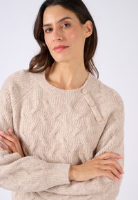Pull en tricot beige avec un col rond et trois boutons décoratifs sur l'épaule gauche. Texture douce avec des poignets à côtes.