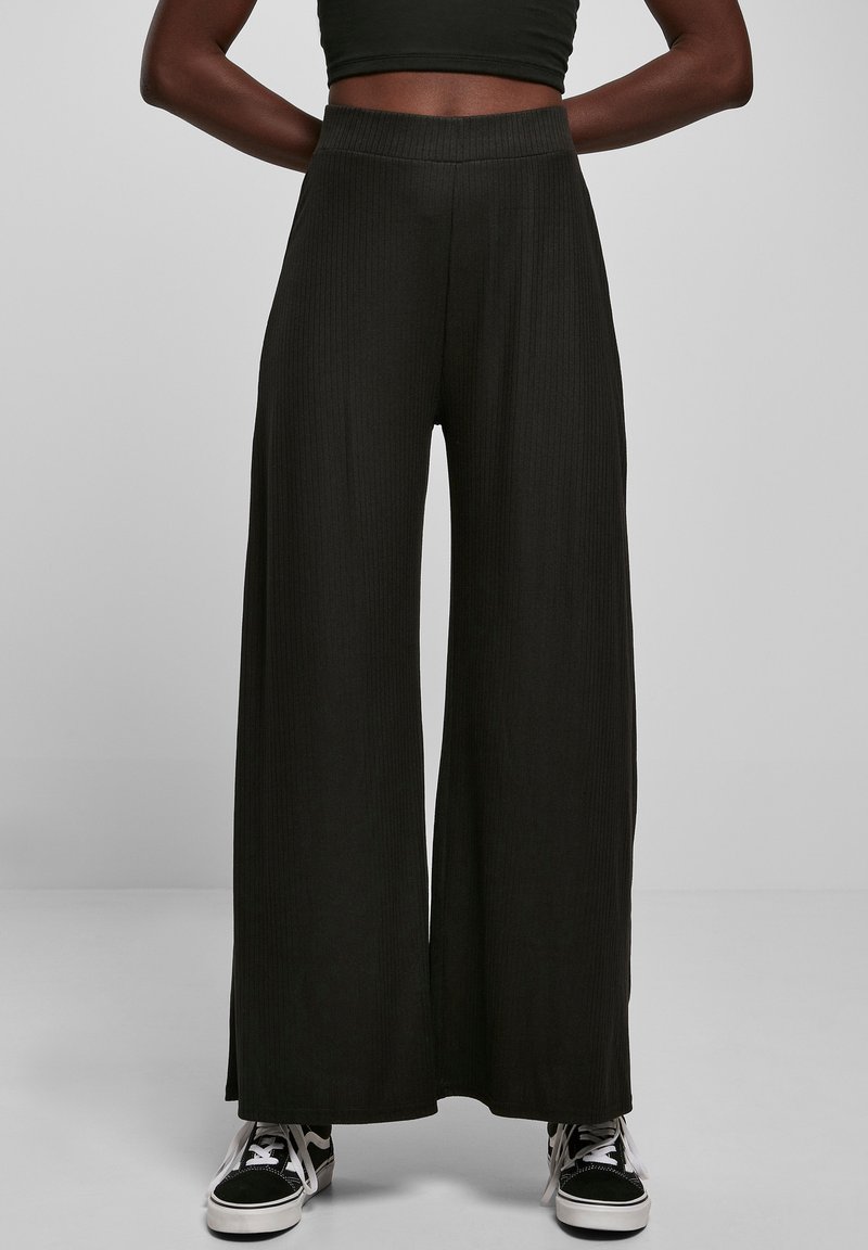 Pantalons larges noirs en tissu côtelé. Taille élastique haute, silhouette fluide, portés avec des baskets noir et blanc.