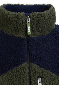 Gros plan d'une veste polaire verte et bleu marine avec un col montant et une fermeture éclair avec une languette verte portant l'inscription "LNRF."