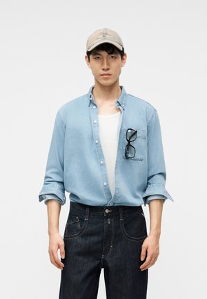 Νέος άνδρας φοράει μπεζ καπέλο, ανοιχτό γαλάζιο τζιν πουκάμισο πάνω από λευκό μπλουζάκι, σκούρο τζιν, με μαύρα γυαλιά κρεμασμένα στην τσέπη του πουκαμίσου.