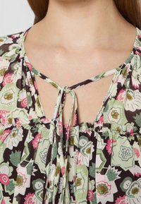 Blumenbluse mit schwarzem Grund und grünen sowie pinken Mustern, einem Schlüssellochausschnitt und Bindebändern an der Vorderseite. Leichter Stoff, gerafftes Design.
