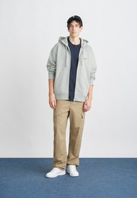 Felpa grigia chiara con zip e cappuccio, indossata sopra una maglietta blu navy, abbinata a pantaloni cargo beige e scarpe da ginnastica bianche. Un cappello scuro completa il look.