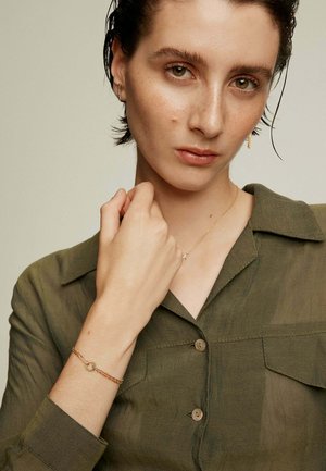 Pulsera de cadena de oro con un acento circular, llevada en una mano de piel clara, combinada con una camisa verde oliva de cuello.