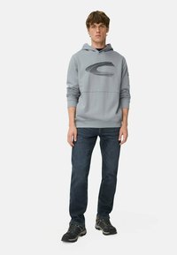 Grijze hoodie met een zwart grafisch logo, gecombineerd met donkerdenim jeans en zwart schoeisel, staande tegen een witte achtergrond.