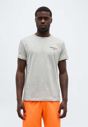 Mees, kes kannab helehallit Björn Borg T-särki ja ereoranžeid lühkesi pükse, seisab neutraalse ilmega valgel taustal.