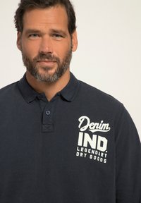 Marinblå pikétröja med krage, tvåknappsstängning och strukturerad tyg. Har vit tryckt text på vänster bröstområde.