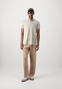 Helles beiges Poloshirt mit kurzen Ärmeln und Kragen, beige Hose und weiße Sneakers mit einer beigen Sohle. Sonnenbrille und braune Mütze.