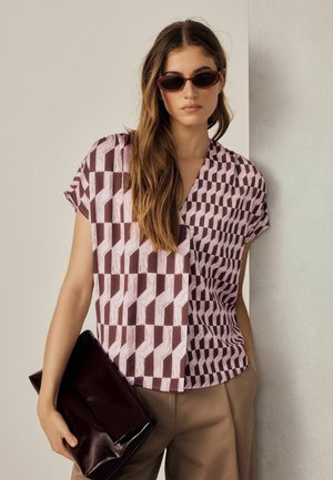 Femme aux longs cheveux bruns portant un haut à motif géométrique et des lunettes de soleil, tenant une grande pochette sombre, debout contre un mur clair.