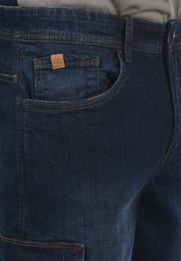 Jeans de mezclilla azul oscuro con un bolsillo trasero que presenta una etiqueta de marca de cuero beige, costuras en contraste y un corte ligeramente ajustado.