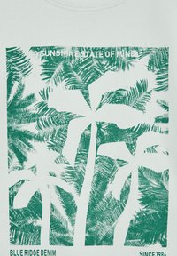 T-shirt bianco con grafica di una palma verde dal tema tropicale, testo "SUNSHINE STATE OF MIND" e informazioni sul marchio in basso.