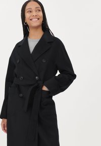 Svart dubbelknäppt kappa med knytbälte, stora framfickor och notchad krage. Tillverkad av strukturerat tyg med slät yta.