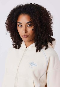 Beige Zip-Hoodie mit blauen Akzenten, mit einem Logo auf der Brust, aus weichem Material gefertigt, mit lockerer Passform und Kängurutasche.