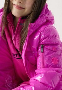 Felroze, glanzend donsjack met capuchon, ritssluitingen aan de zakken en een logopatch. Daaronder is een bijpassende geribbelde roze trui zichtbaar.