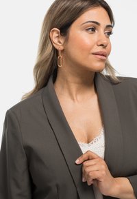 Blazer grigio con colletto sciallato, abbinato a un top in pizzo bianco. Presenta orecchini a cerchio in oro rosa e capelli lisci e texturizzati.
