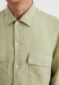 Chemise en lin vert clair avec un col, deux poches poitrine et boutons beiges. Texture visible avec des détails de couture.