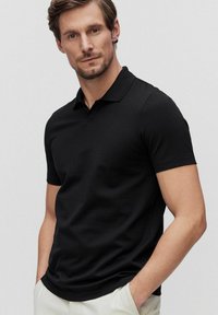 Svart kortärmad polo med en mjuk yta, klassisk krage och en åtsittande design, kompletterad med subtil branding vid fållen.