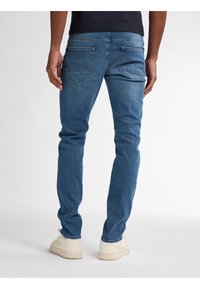 Vaqueros de mezclilla en azul medio con un diseño ajustado, que presentan bolsillos traseros con un patrón de costura único. Llevados con zapatillas de deporte en color blanco roto.