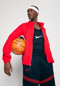 Rød atletisk jakke over en sort tanktop, holder en orange basketball. Sorte shorts med en rød stribe og logo-detail. Hvidt pandebånd båret.