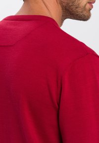 Czerwony sweter z okrągłym dekoltem, z ribbowanymi detalami przy kołnierzu i mankietach, o gładkiej, miękkiej fakturze. Zbliżenie z tyłu.