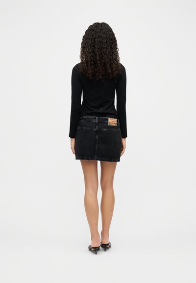 Mini-jupe en denim noir avec taille ajustée, coupe droite et détail d'étiquette à l'arrière, associée à un haut noir à manches longues et des talons.
