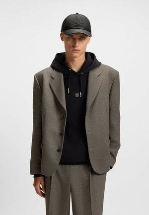 Brown textured blazer with black buttons over a black hoodie. Model wears a black cap. The ensemble features a relaxed fit.