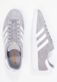 Szare zamszowe trampki z trzema białymi paskami, teksturowaną podeszwą i złotymi detalami z logo na języku. Klasyczny niskoprofilowy design.